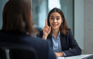 Giovane ragazza durante un colloquio di lavoro che solleva con garbo l'indice per chiedere di porre una domanda alla selezionatrice di fronte a lei.