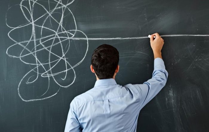 Uomo in camicia visto di spalle mentre traccia con il gesso una linea bianca retta e decisa su una lavagna, facendola emergere da un ammasso confuso di linee e scarabocchi.