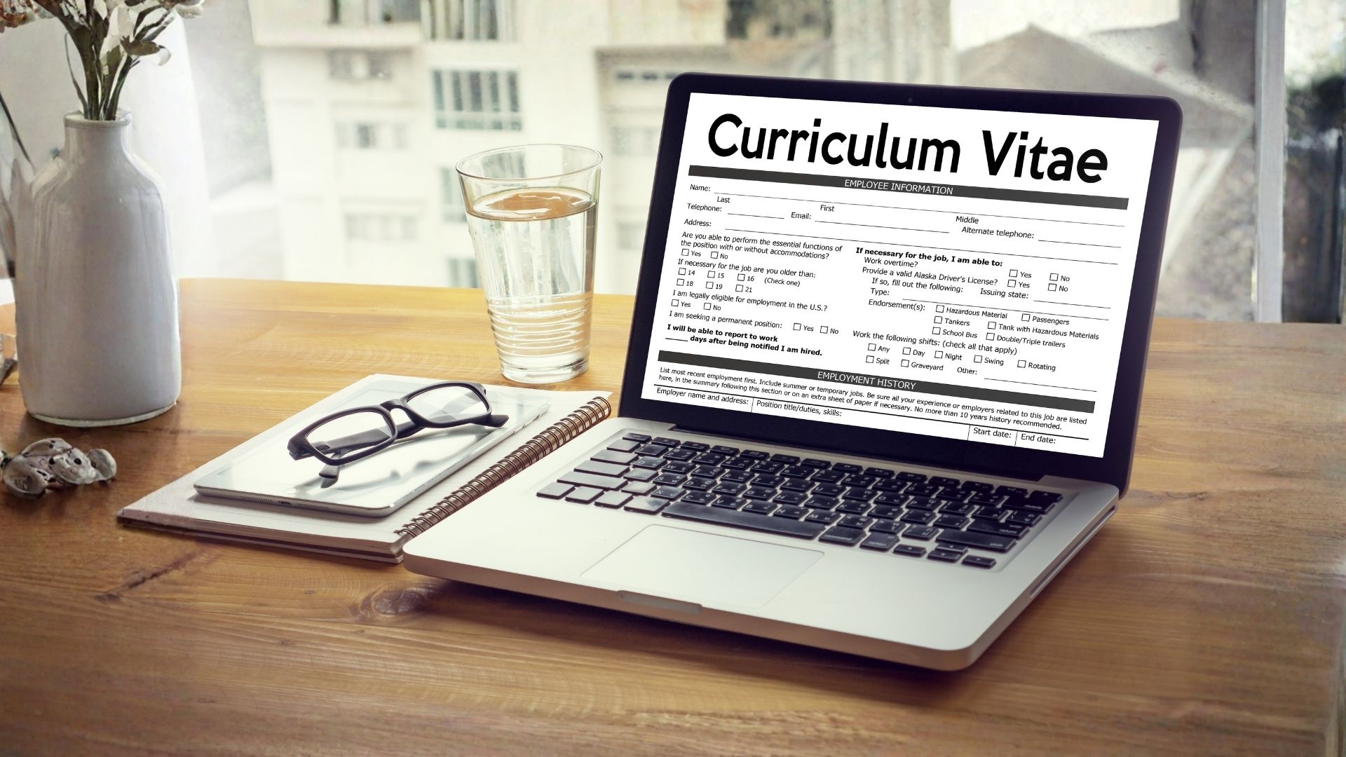 Postazione di lavoro minimalista: computer con curriculum, occhiali e agenda su un tavolo pulito.