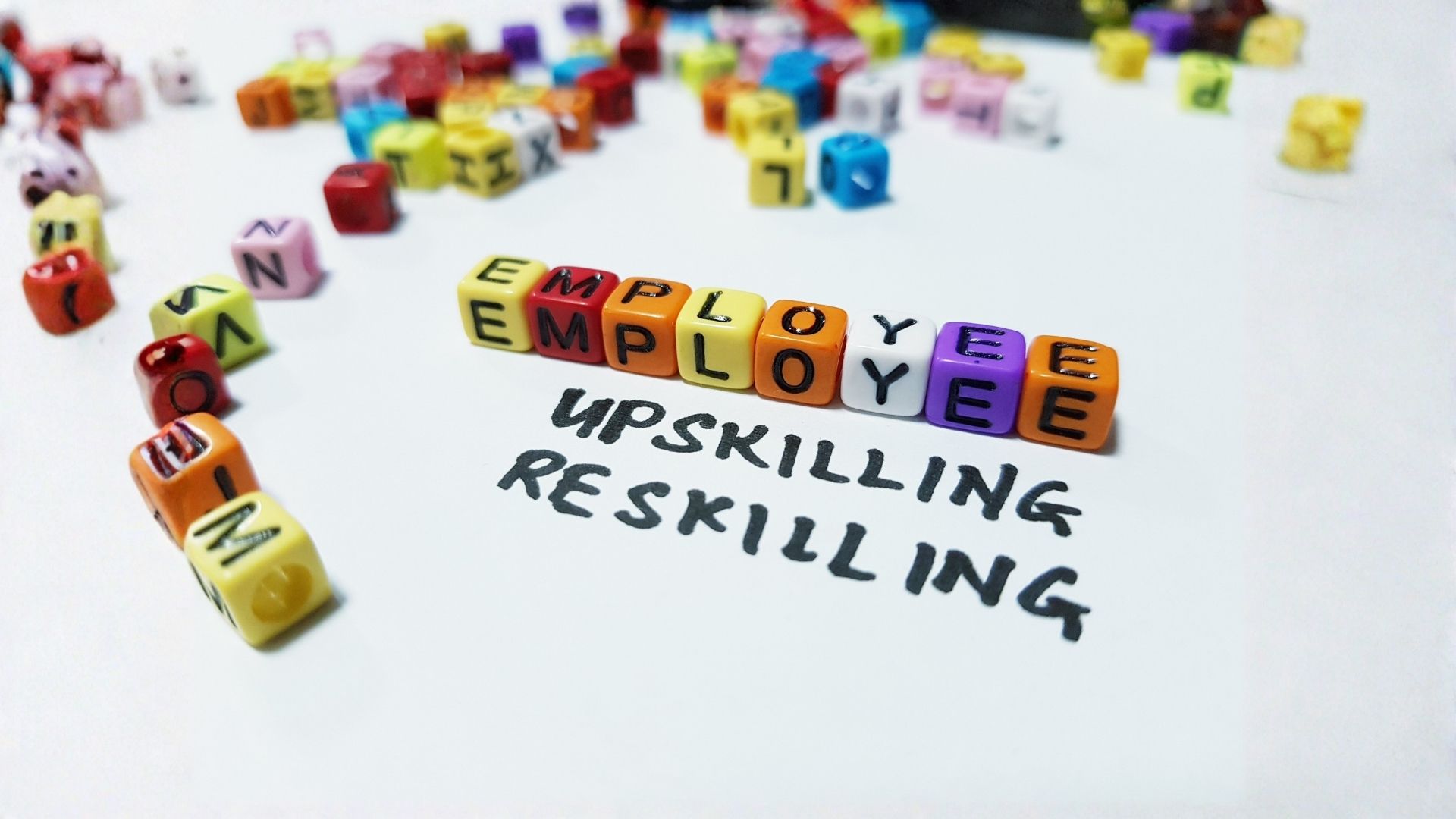 Primo piano di cubetti colorati che compongono la parola "EMPLOYEE" sopra le scritte a mano "UPSKILLING" e "RESKILLING" su un foglio bianco.