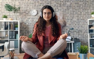Una giovane donna seduta sulla scrivania di un ufficio moderno in posizione di meditazione (loto). Indossa delle cuffie, ha gli occhi chiusi e un'espressione serena, circondata da un ambiente di lavoro luminoso.