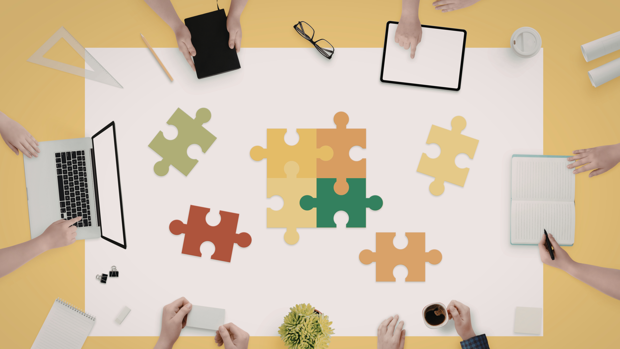 Immagine di mani di diverse persone riunite attorno a un tavolo di lavoro, come in un team: alcune sono su tastiere di computer, altre vicine a una tazza di caffè. Al centro del tavolo ci sono pezzi di puzzle sparsi che rappresentano il concetto di job crafting, simbolo di costruzione e personalizzazione del proprio lavoro.